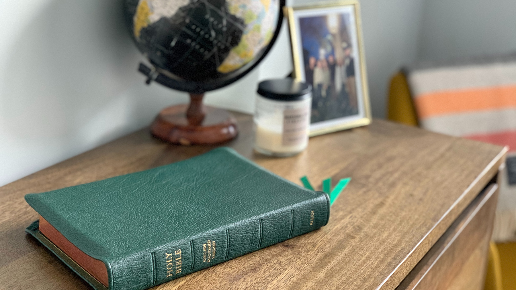 global professionals_tim-wildsmith-5Pcmc8LLVQs-unsplash Green ESV Bible on a desk with a globe, candle, and family photo in the background.