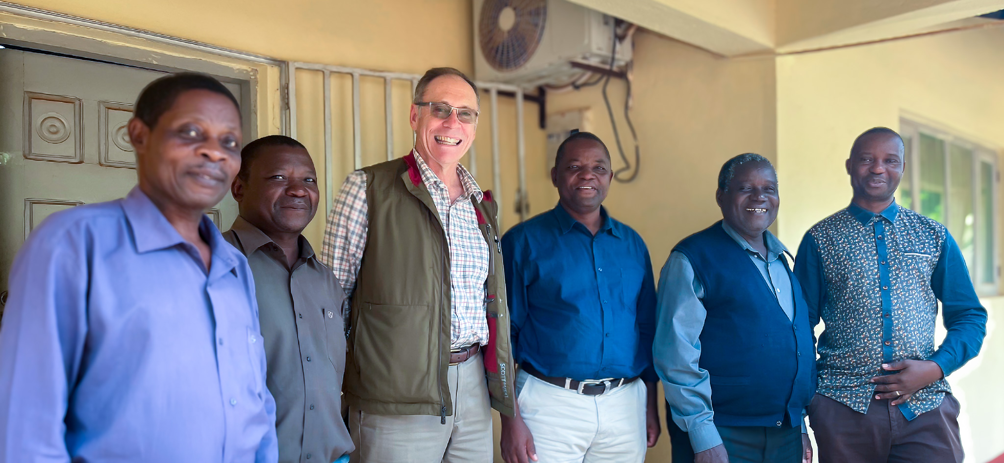 Stuart Foster-2 A group of six men standing side by side in front of a yellow building, smiling at the camera. One man in the center is wearing glasses, a patterned shirt, and a vest, while the others are dressed in collared shirts and slacks. An air conditioning unit is visible on the wall above them.